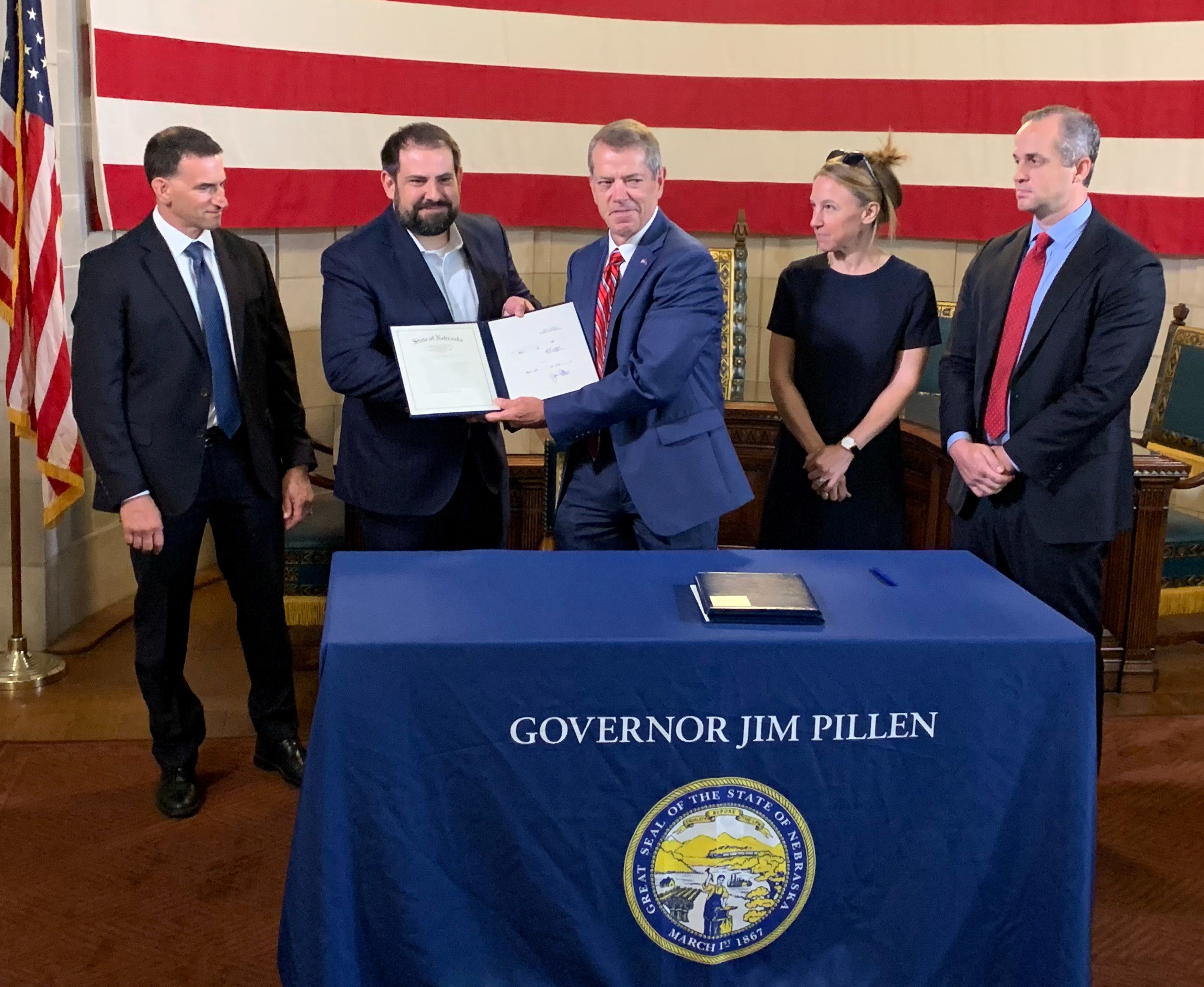 Gov. Pillen presents a copy of LB 1096 to Senator Bostar while standing behind a table with a blue table cloth that says "Governor Jim Pillen" above an image of the Great Seal of the State of Nebraska. The original copy of the bill lies on top of the table. FBI Special Agent Eugene Kowel stands to their right. Jackie Deal, with State Armor and founder, CEO, and Chairman of State Armor Michael Lucci stand to their left.