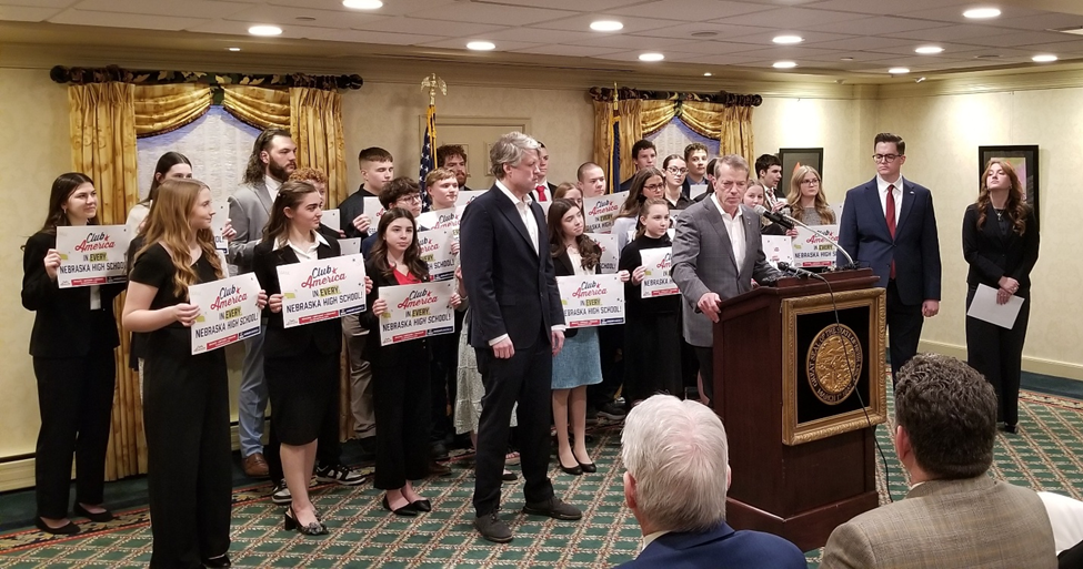 Governor Pillen, with Attorney General Mike Hilgers standing to his right, Nick Cocca, TPUSA Club America Enterprise Director and Chapter President Emma Smith, of Creighton University standing to his left, and students standing behind him holding signs saying "Club America in every Nebraska High School", addresses the audience.