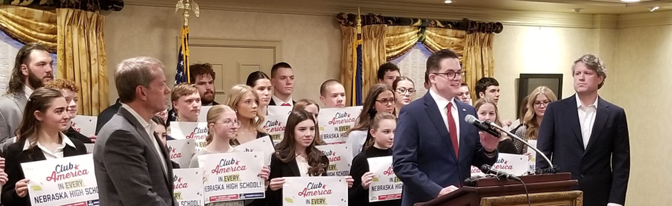 Nick Cocca, TPUSA Club America Enterprise Director and Chapter President, with Governor Pillen standing to his right, Attorney General Mike Hilgers standing to his left, and students standing behind him holding signs saying "Club America in every Nebraska High School", addresses the audience.