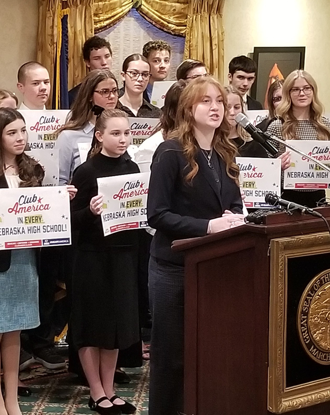 Chapter President Emma Smith, of Creighton University with students standing behind her holding signs saying "Club America in every Nebraska High School", addresses the audience.