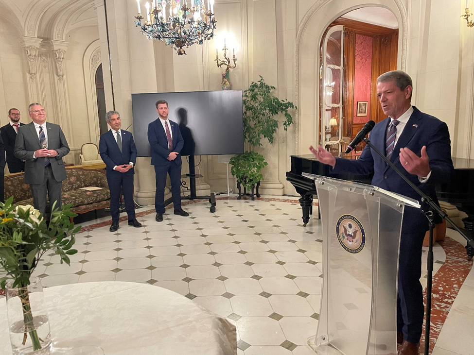 Gov. Pillen speaks at reception at the residence of U.S. Ambassador Bijan Sabet. Sabet is second to the left, standing at the television screen.