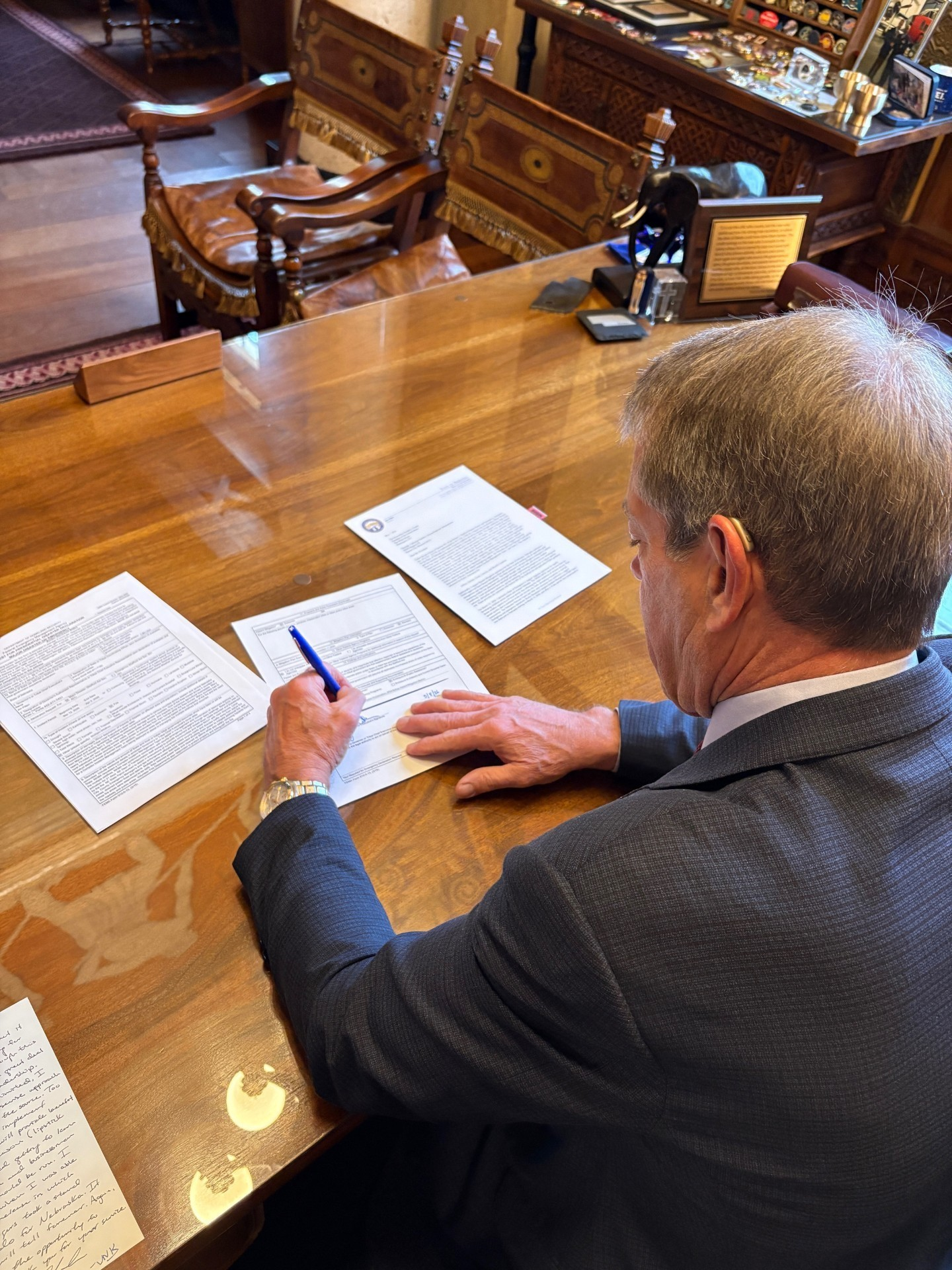 Image from behind Gov. Pillen as he sits at his desk signing a request for Federal Disaster Declaration for Wildfire Damage