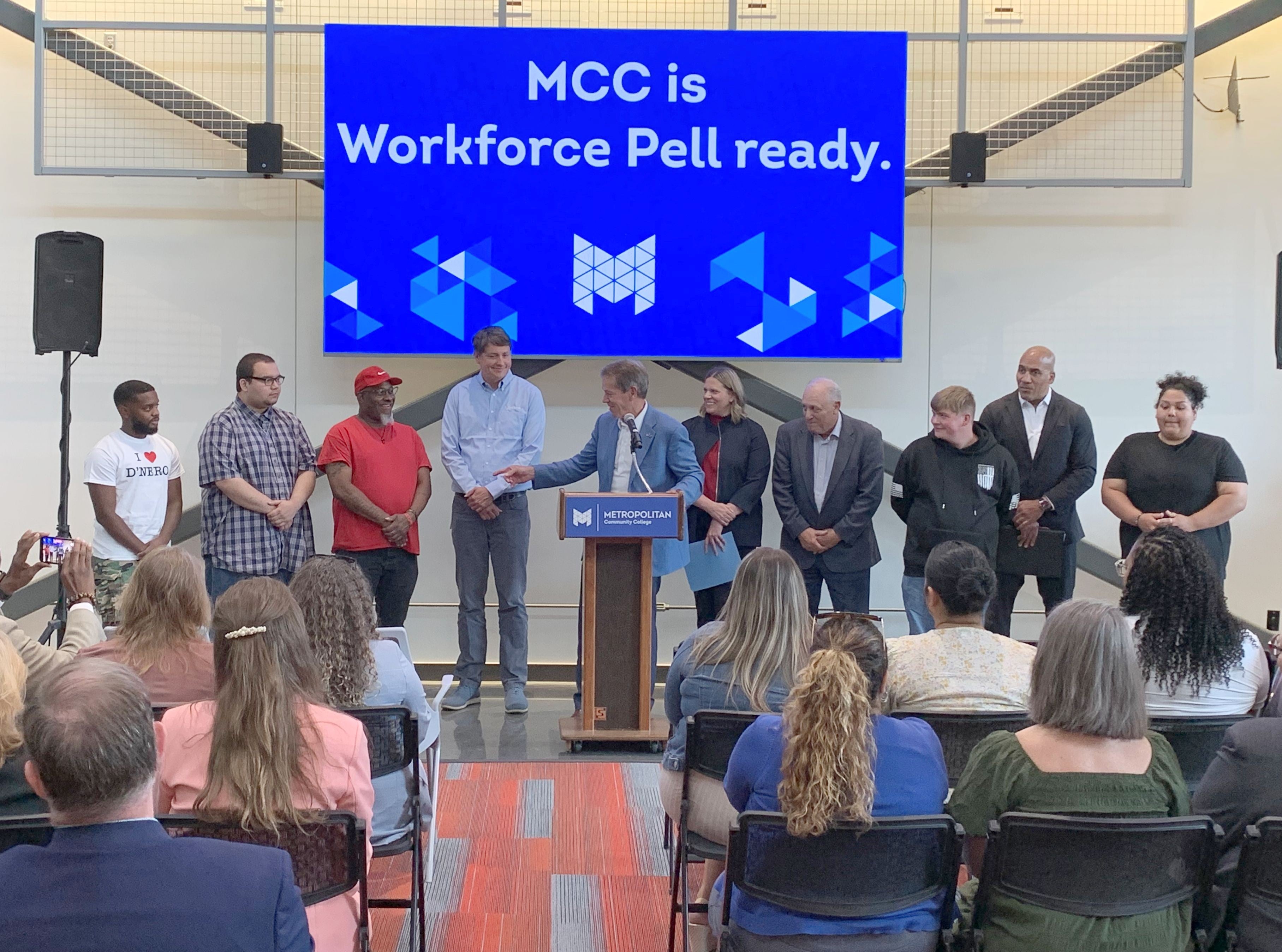 Governor Jim Pillen speaks from a podium at Workforce Pell Grant announcement as an audience listens. He is flanked by students, Andy Lozier, CEO at Lozier Corporation, NDOL Commissioner Katie Thurber, Randy Schmailzl, Metropolitan Community College president, and Rob Jeffreys, director of the Department of Corrections. A sign behind them says, "MCC is Workforce Pell ready."