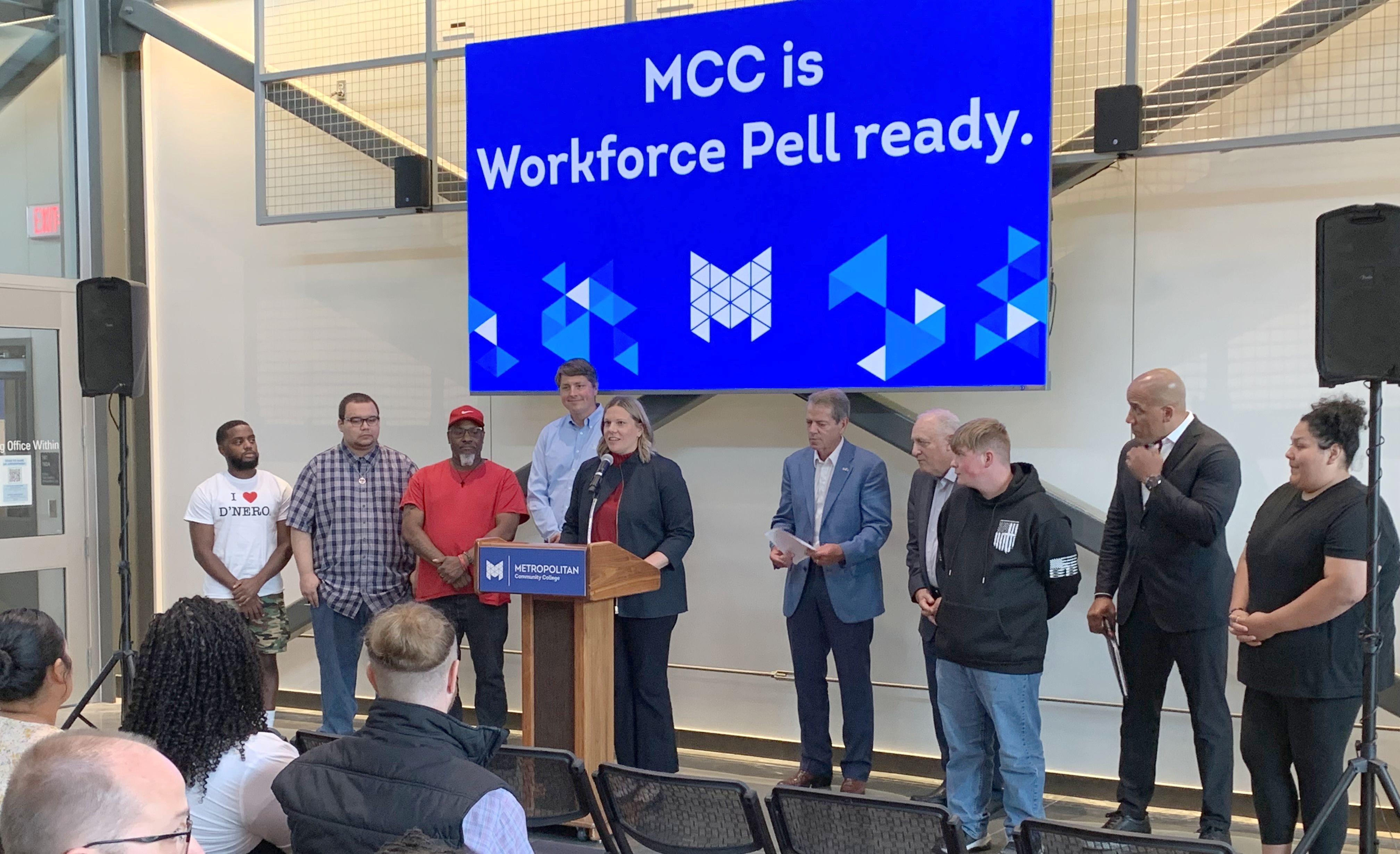 NDOL Commissioner Katie Thurber speaks from a podium at Workforce Pell Grant announcement as an audience listens. She is flanked by students, Andy Lozier, CEO at Lozier Corporation, Governor Jim Pillen, Randy Schmailzl, Metropolitan Community College president, and Rob Jeffreys, director of the Department of Corrections. A sign behind them says, "MCC is Workforce Pell ready."