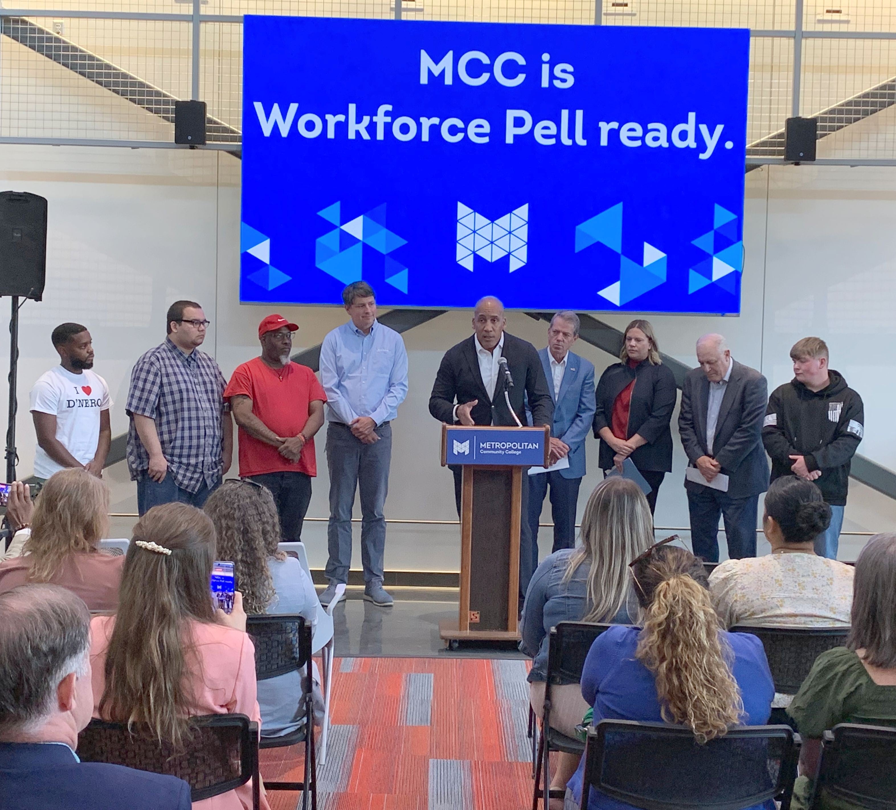 Rob Jeffreys, director of the Department of Corrections speaks from a podium at Workforce Pell Grant announcement as an audience listens. He is flanked by students, Andy Lozier, CEO at Lozier Corporation, Governor Jim Pillen, NDOL Commissioner Katie Thurber, and Randy Schmailzl, Metropolitan Community College president. A sign behind them says, "MCC is Workforce Pell ready."