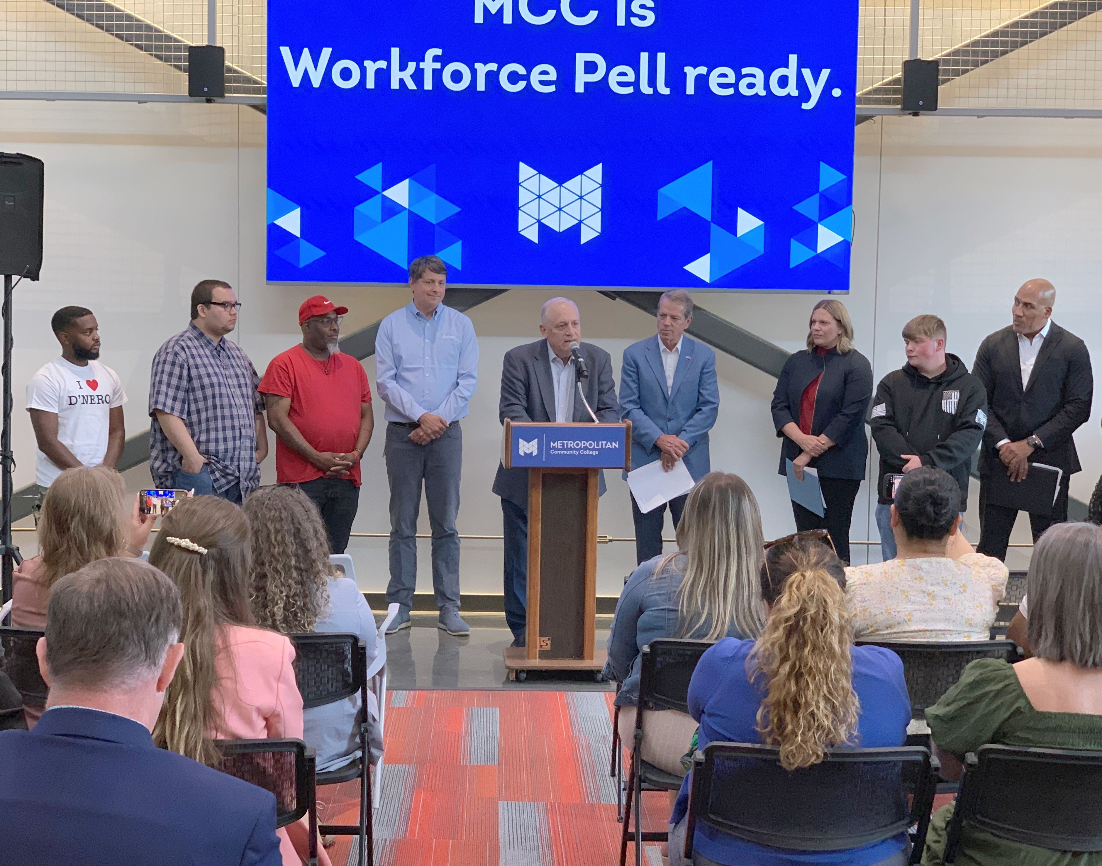Randy Schmailzl, Metropolitan Community College president speaks from a podium at Workforce Pell Grant announcement as an audience listens. He is flanked by students, Andy Lozier, CEO at Lozier Corporation, Governor Jim Pillen, NDOL Commissioner Katie Thurber, and Rob Jeffreys, director of the Department of Corrections. A sign behind them says, "MCC is Workforce Pell ready."