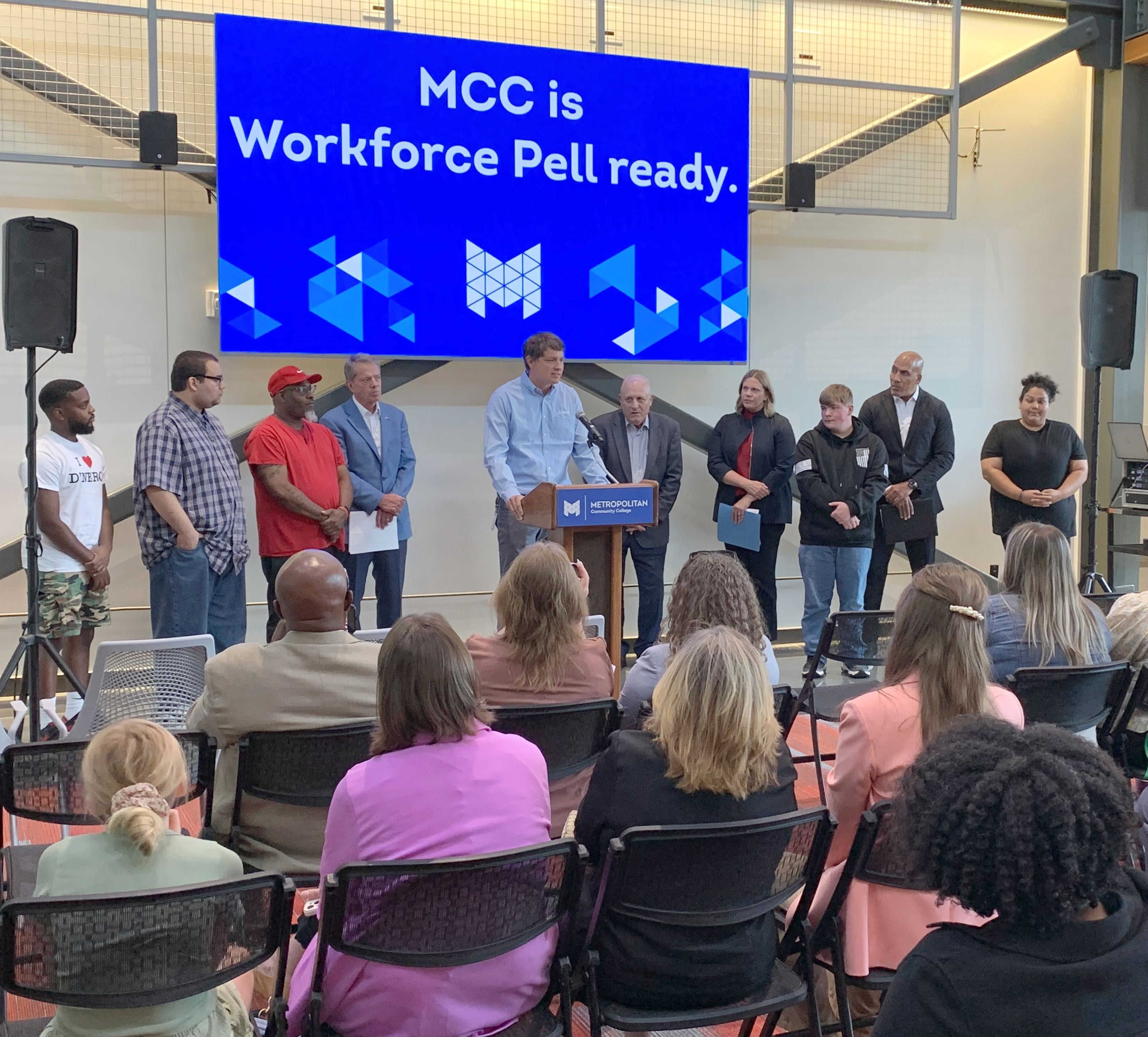 Andy Lozier, CEO at Lozier Corporation speaks from a podium at Workforce Pell Grant announcement as an audience listens. He is flanked by students, Governor Jim Pillen, Randy Schmailzl, Metropolitan Community College president, NDOL Commissioner Katie Thurber, and Rob Jeffreys, director of the Department of Corrections. A sign behind them says, "MCC is Workforce Pell ready."
