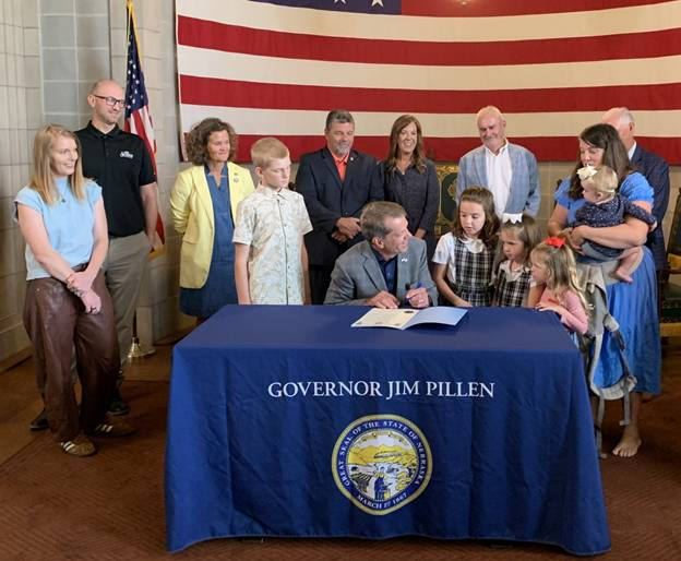 Governor Pillen, surrounded by some students, with Senators and other parents behind them, signs the proclamation.