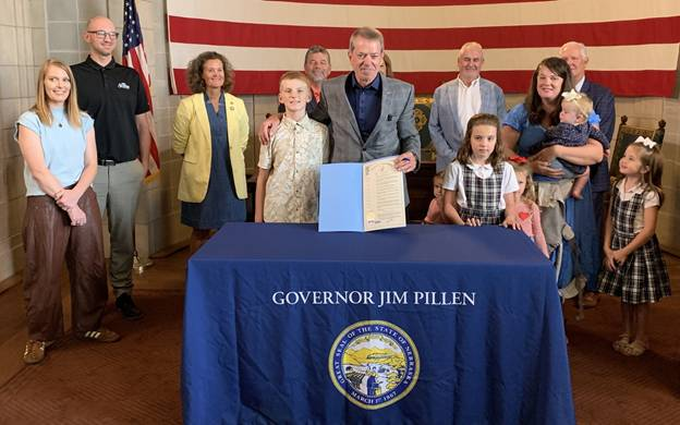 Governor Pillen, surrounded by some students, with Senators and other parents behind them, displays the proclamation for the audience.
