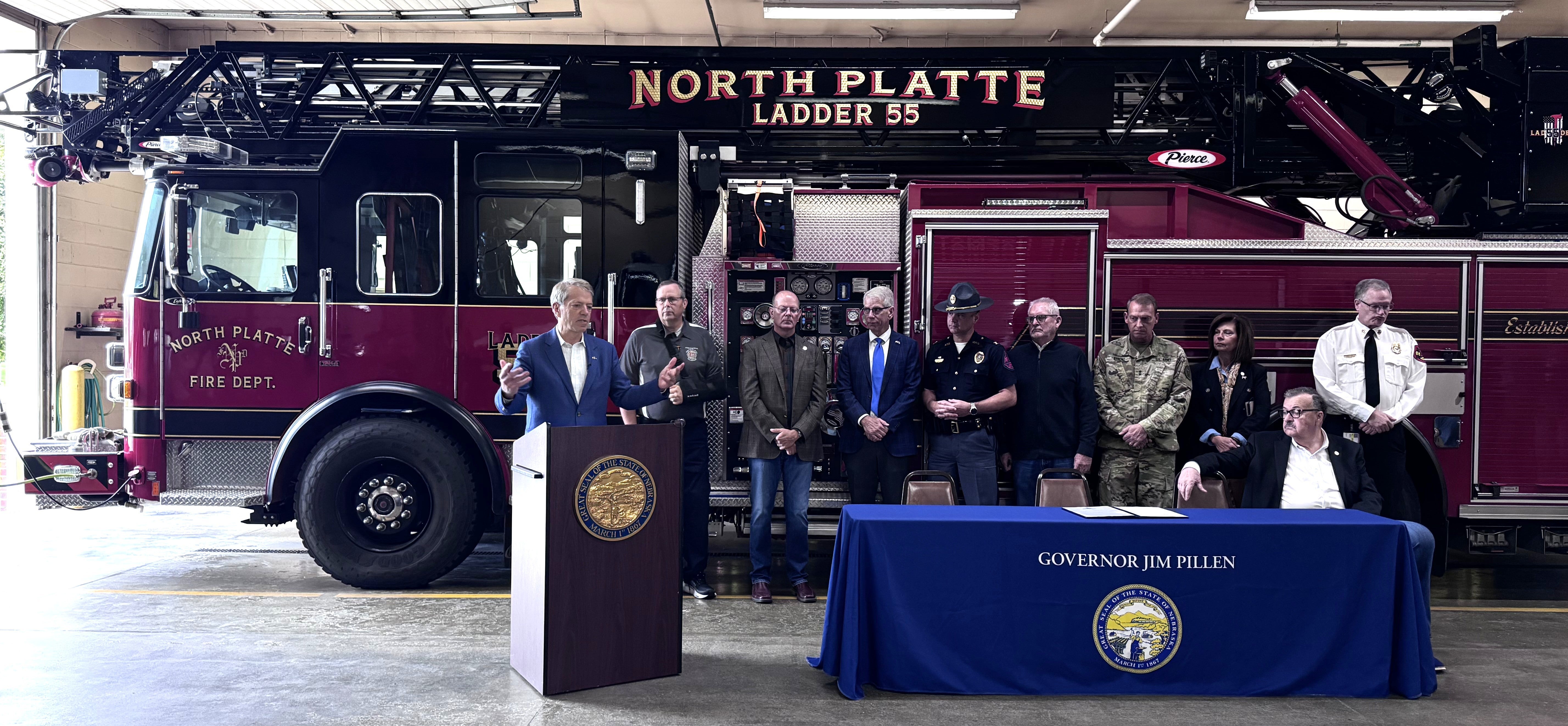 Governor Jim Pillen addressed an audience at North Platte Fire Station 3 from a podium. He is joined by members of the new task force. A table with a covering that says Governor Jim Pillen with the Great Seal of the State of Nebraska on it is in front of them. Behind them is North Platte Ladder 55 fire truck.