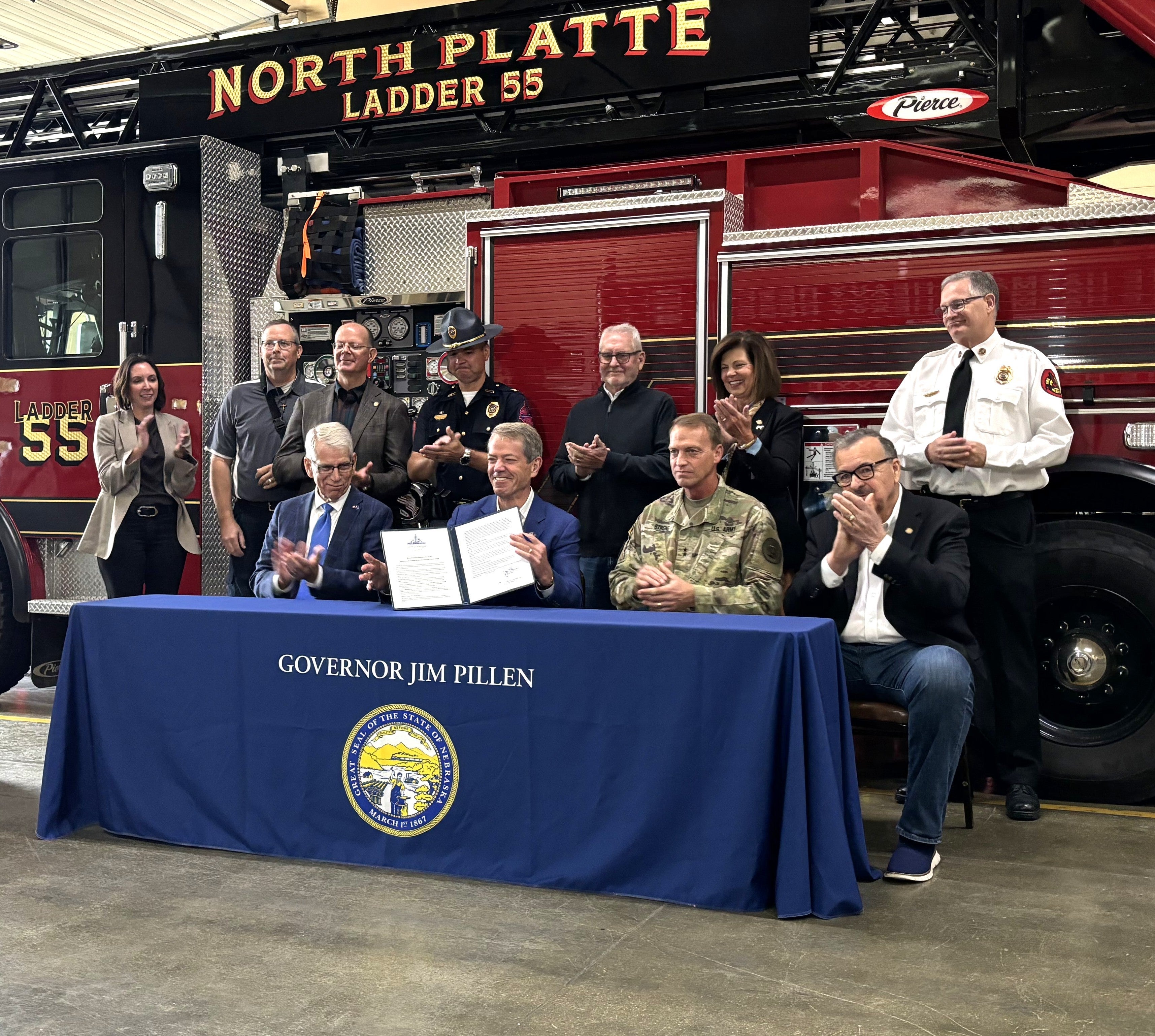 Governor Jim Pillen displays the signed executive order while seated behind a table with a covering that says Governor Jim Pillen with the Great Seal of the State of Nebraska on it. Members of the new task force are seated beside him, with some standing behind them. North Platte Ladder 55 fire truck is behind everyone.