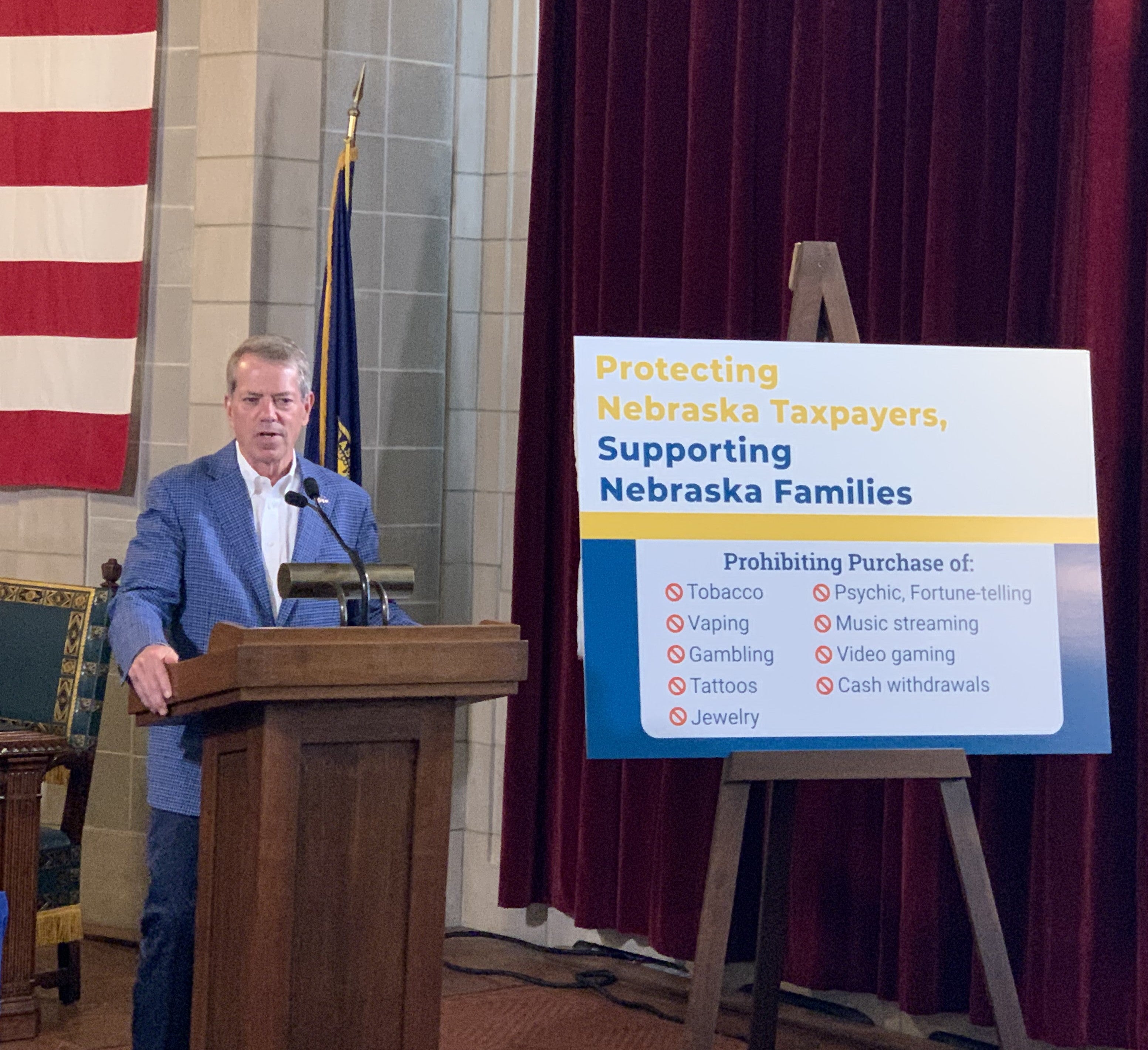 Governor Jim Pillen addresses the audience on executive order restricting TANF funds. Beside him a sign reads: "Protecting Nebraska Taxpayers, Supporting Nebraska Families. Prohibiting Purchase of: Tobacco; Vaping; Gambling; Tattoos; Jewelry; Psychic, Fortune-telling; Music streaming; Video gaming; Case withdrawls.