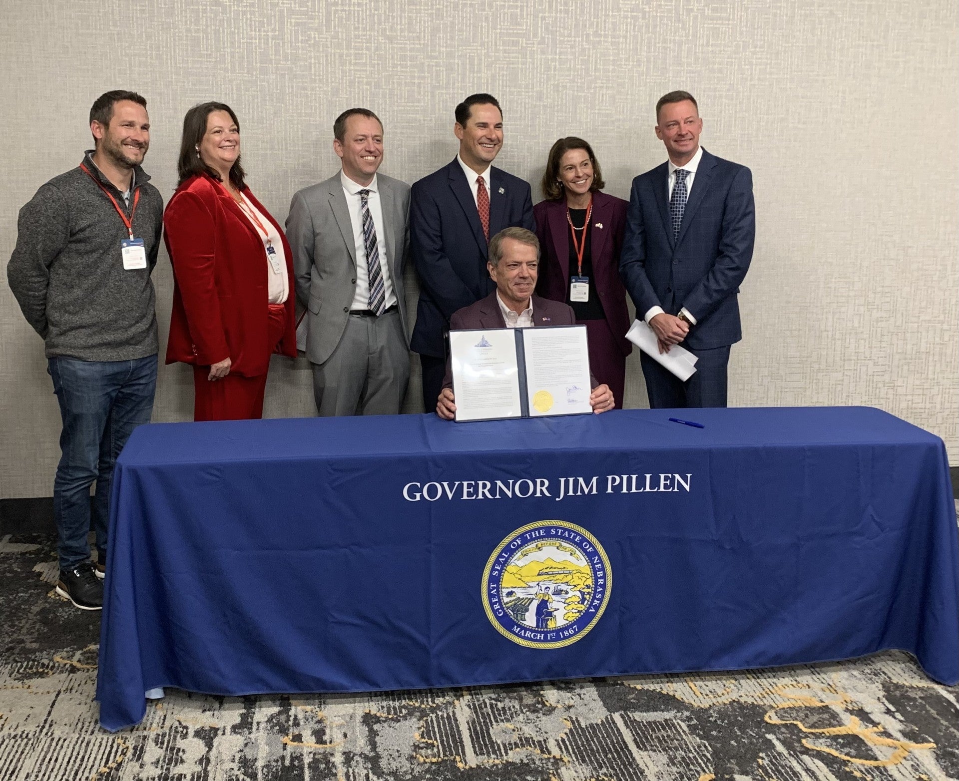 Governor Pillen, which sitting at a table with Governor Jim Pillen in capital letters and an image of the Great Seal of Nebraska, displays the signed copy of Executive Order 26-22 during the Governor's Summit at the Younes Conference Center. Behind him are members of the Governor’s Economic Growth Working Group, chaired by DED Director Maureen Larsen.